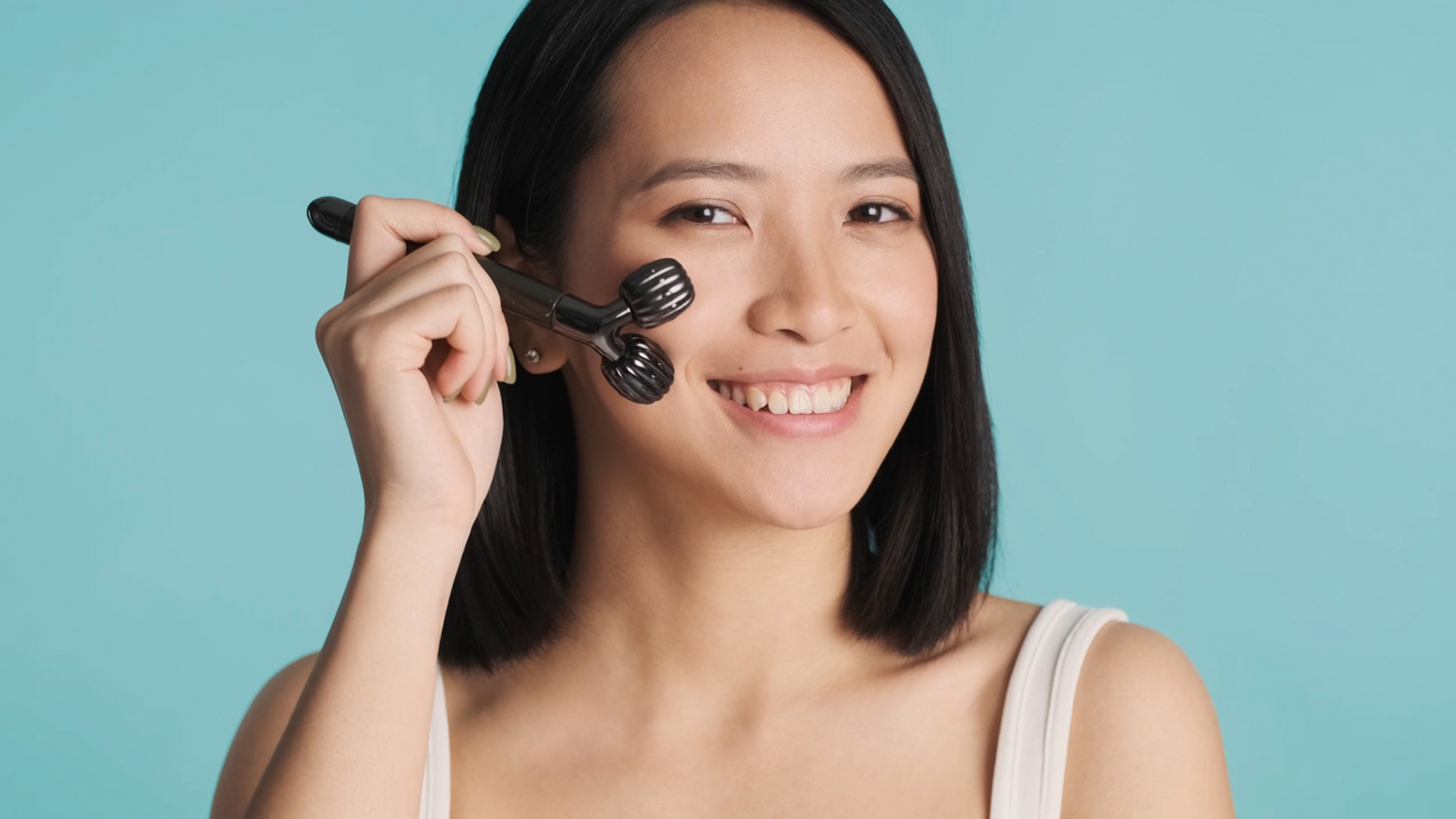 Close up Asian woman performing morning skincare routine massaging face with roller smiling at camera over blue background